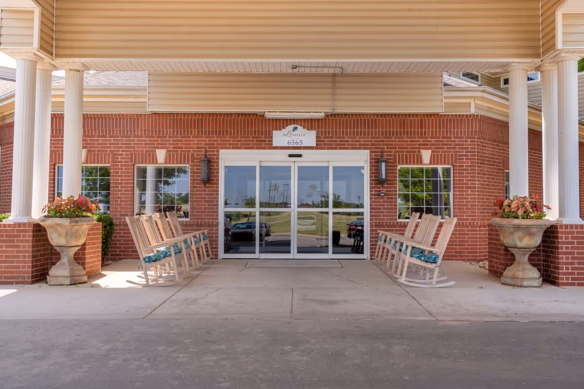 The covered front entrance of a senior living building with brick facade, glass sliding doors flanked by rocking chairs and large planters.