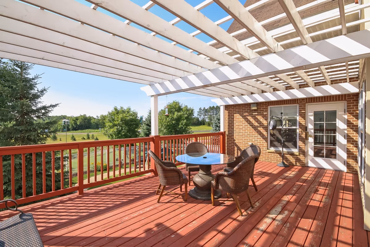 Outdoor wooden deck area with a white pergola overhead, featuring a round glass table surrounded by four wicker chairs. The deck has a red railing and overlooks a green landscape with trees and a clear blue sky. A brick wall with a window and a door is visible on one side of the deck.