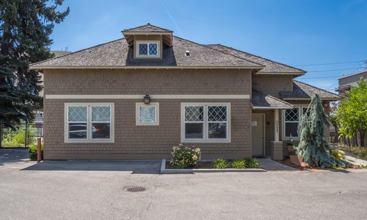 Brown, shingled building exterior with windows, a doorway, and surrounding shrubs.