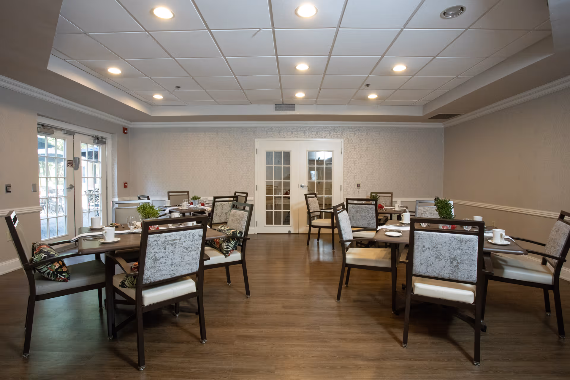 A dining room with several tables and chairs arranged neatly. Each table has place settings including cups, plates, and small plants as centerpieces. The room has wooden flooring, light-colored walls, a drop ceiling with recessed lighting, and French doors at the far end.