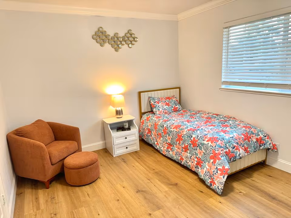 Cozy bedroom featuring a single bed with floral quilt, nightstand, lamp, wood floor, and seating.