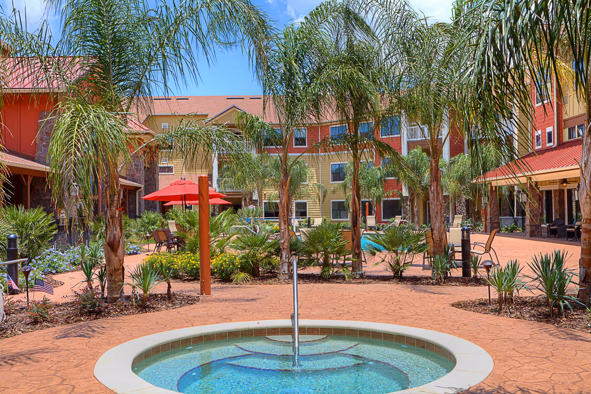 Sunny landscaped courtyard with a circular fountain, palm trees, patio seating, and a surrounding multi-story building.