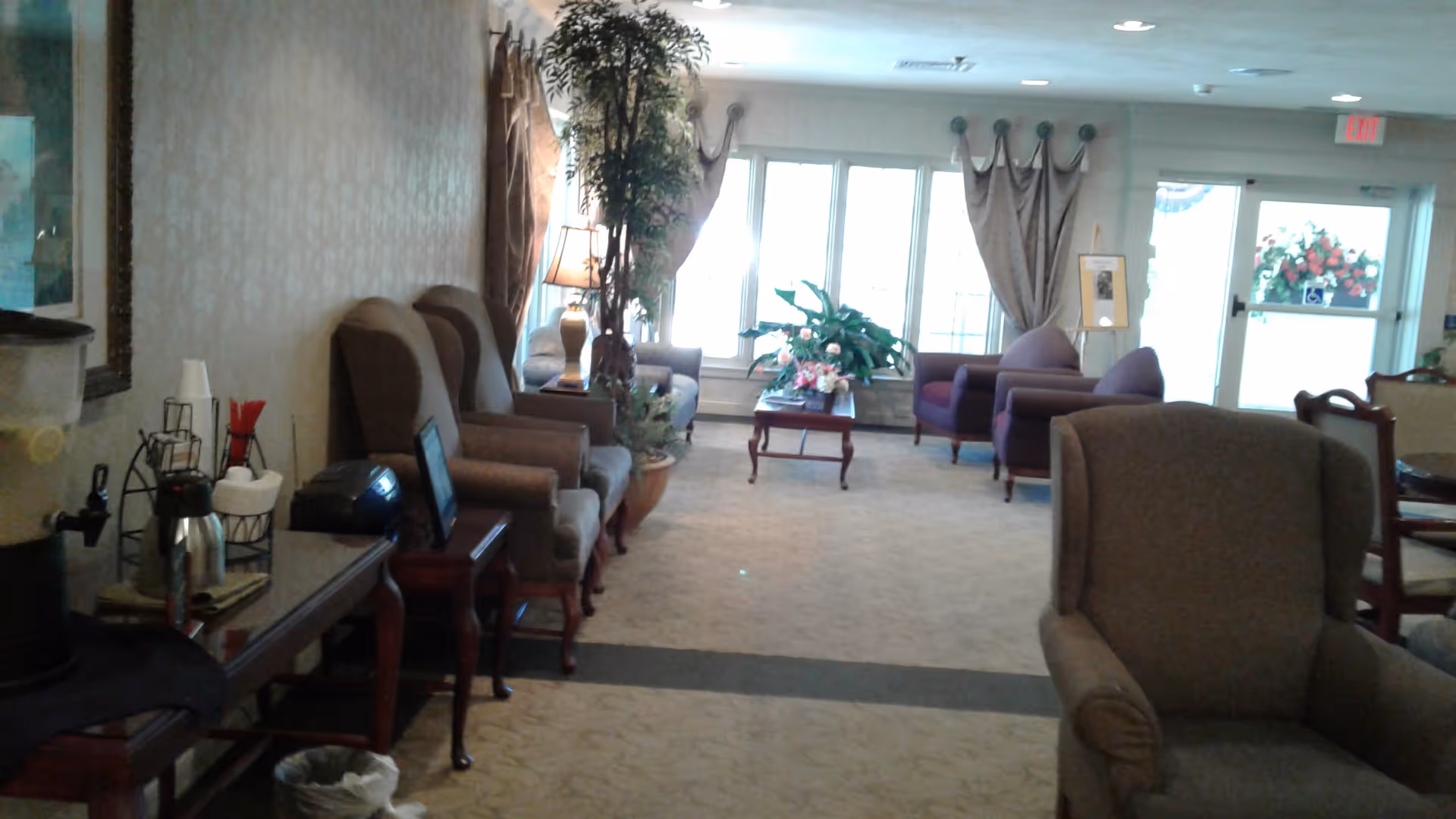 Bright senior living lounge with arranged chairs, sofas, plants, and a coffee station.