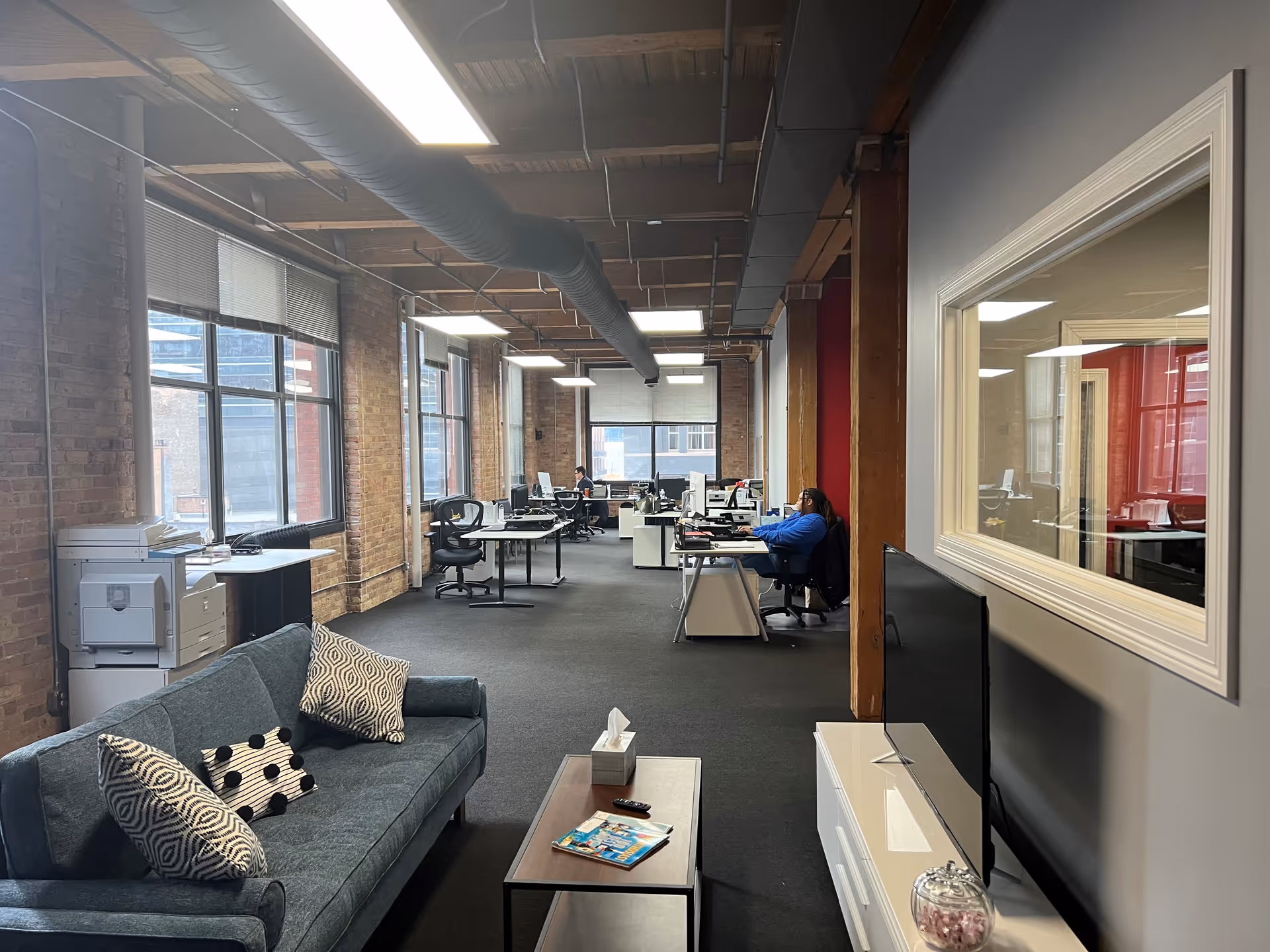 Office-style lounge and open workspace with brick walls, exposed ductwork, and desks with people working.