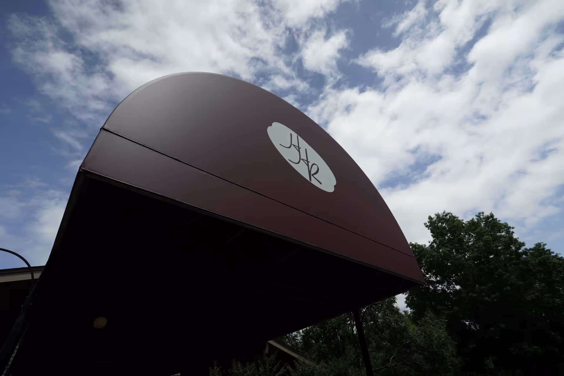 Curved maroon entry canopy with a circular logo, set against blue sky and trees.