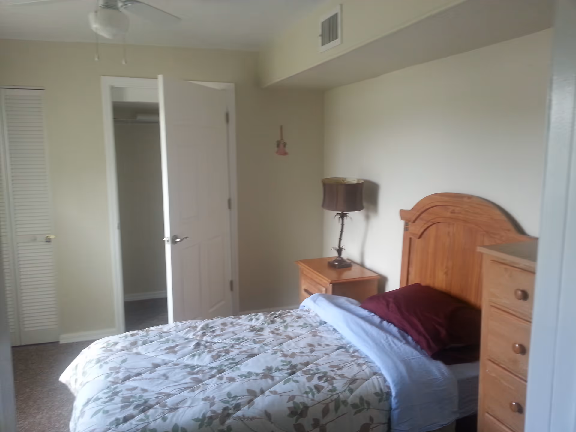A bedroom with a wooden headboard bed covered with a floral patterned comforter and a burgundy pillow. Next to the bed is a wooden nightstand with a lamp, and a wooden dresser is visible on the right side. The room has light-colored walls, a ceiling fan, and an open door leading to a closet.
