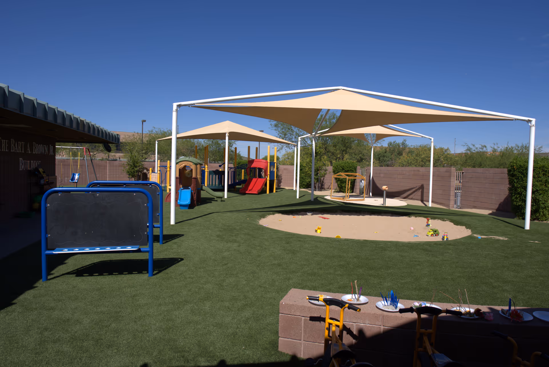 Outdoor play area with shade sails, colorful playground equipment, and sandbox near the facility building.