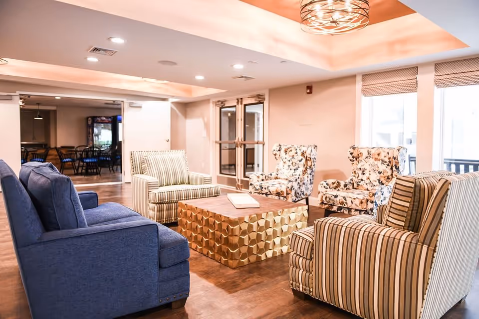 A bright and cozy living room area with a mix of patterned and solid upholstered armchairs arranged around a textured gold coffee table. The room features large windows with blinds, recessed lighting, and a decorative ceiling light fixture. In the background, there is a dining area with tables and chairs.