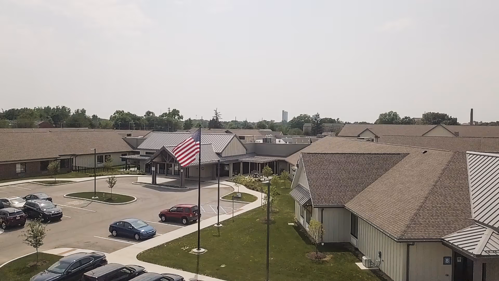 A senior living campus exterior with low-rise buildings, a parking lot, and an American flag.