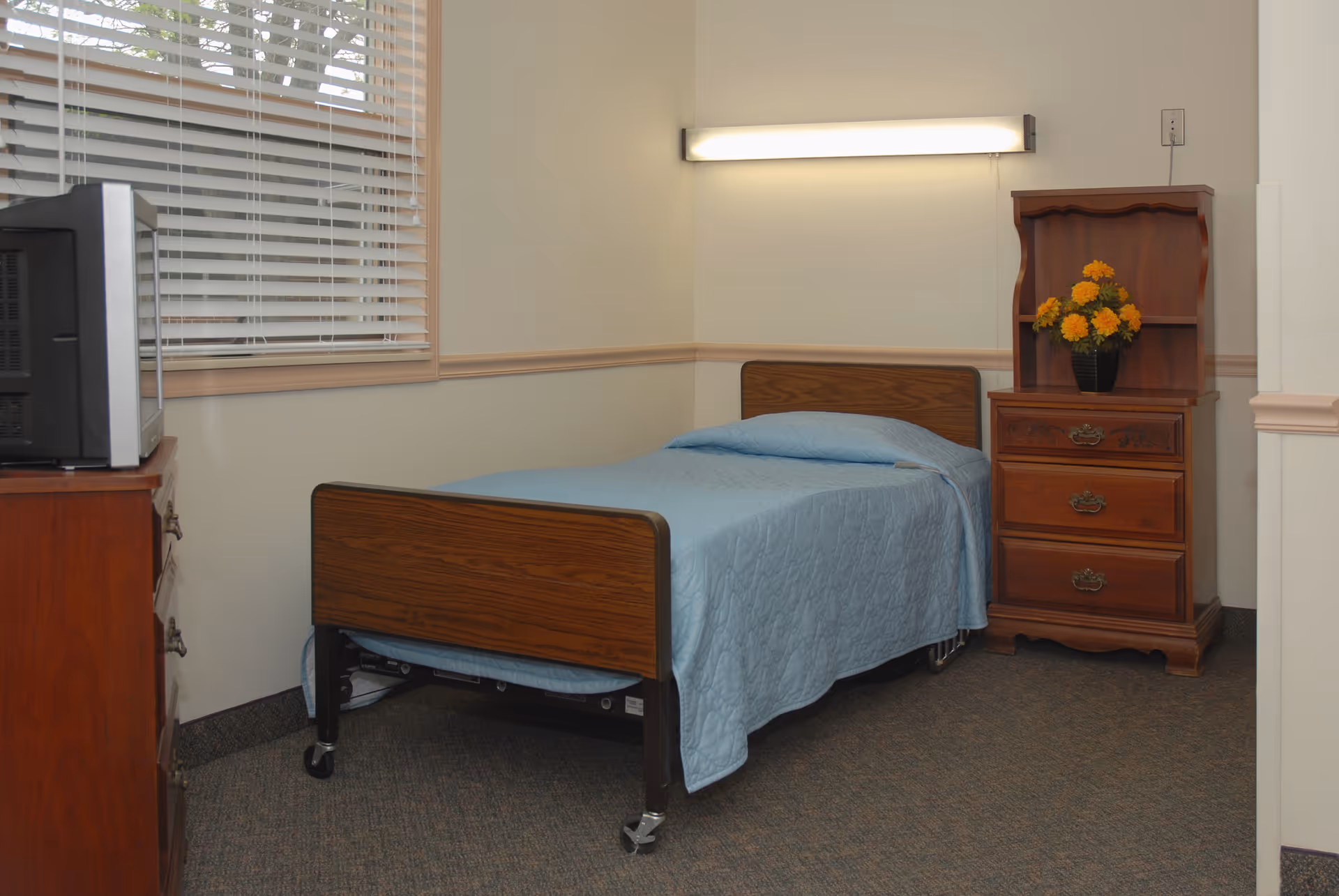 A small bedroom with a single bed covered in a blue bedspread, a wooden dresser with flowers, and a television on a chest.