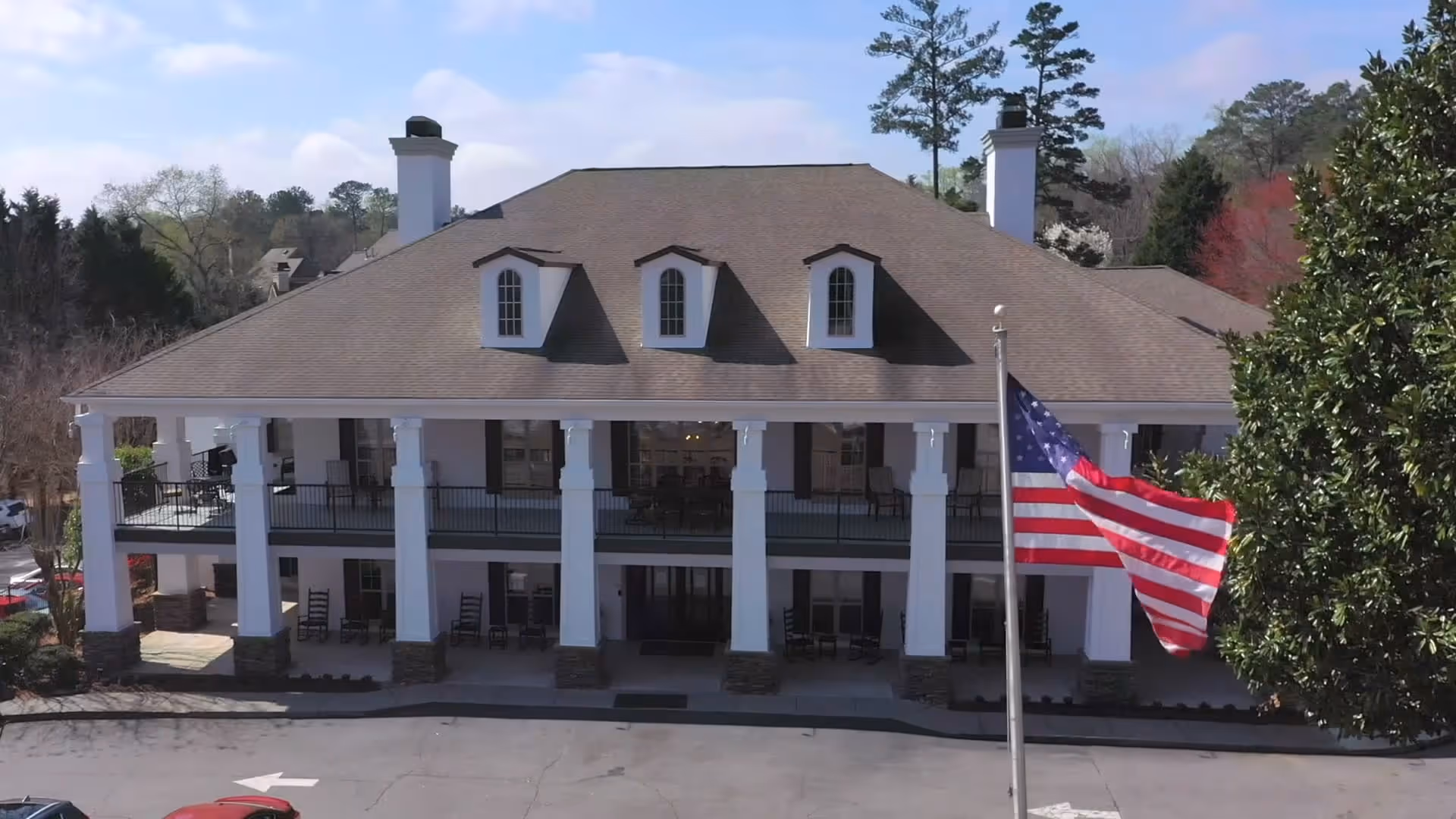 Front exterior view of a large two-story building with white columns and a brown roof, featuring an American flag on a flagpole in front and surrounded by trees.