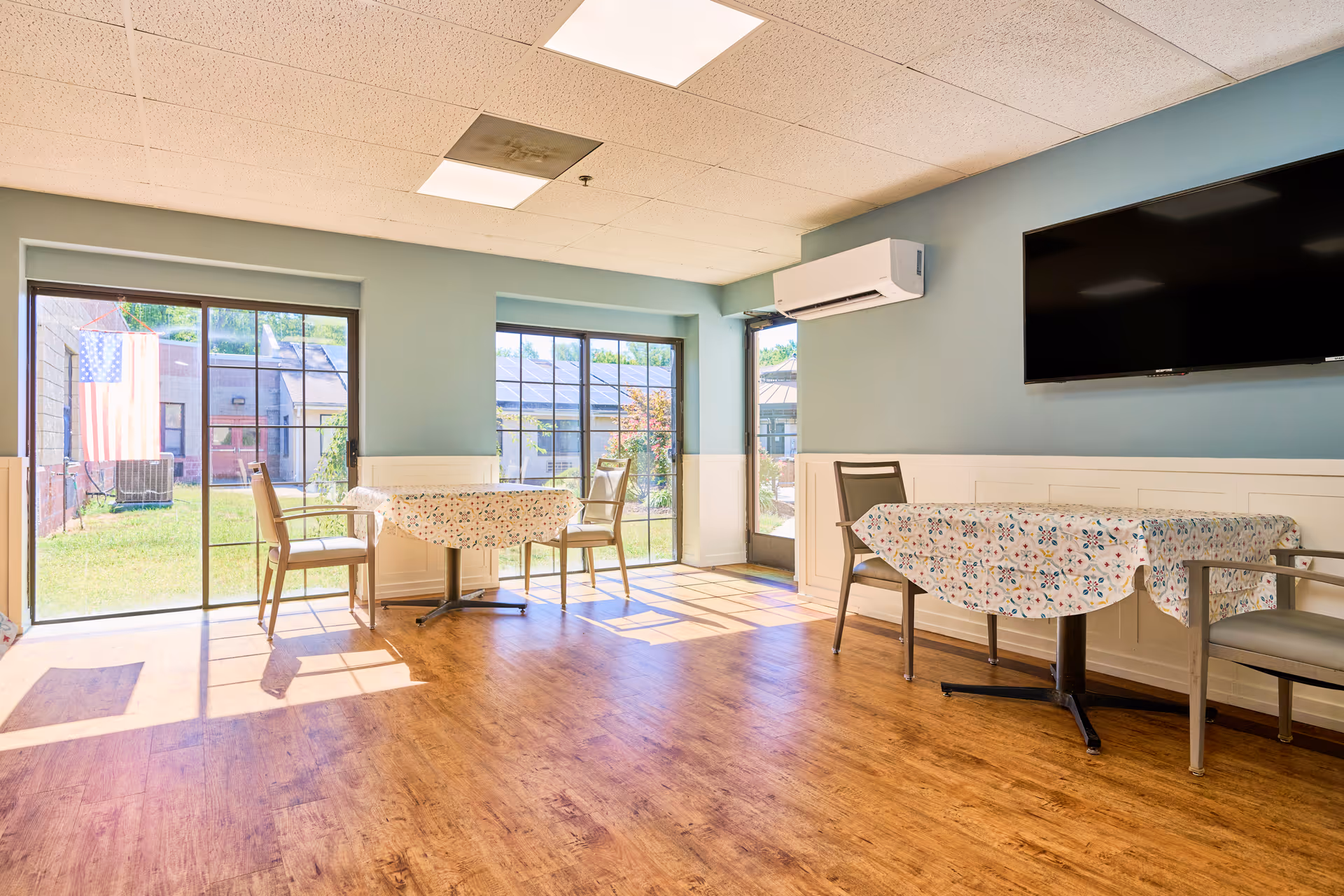 A bright dining room with floral tablecloths, chairs, large windows, and a wall-mounted TV.