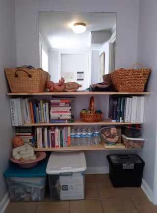 Shelving unit with books, baskets, dolls, water bottles, and bins in a hallway.