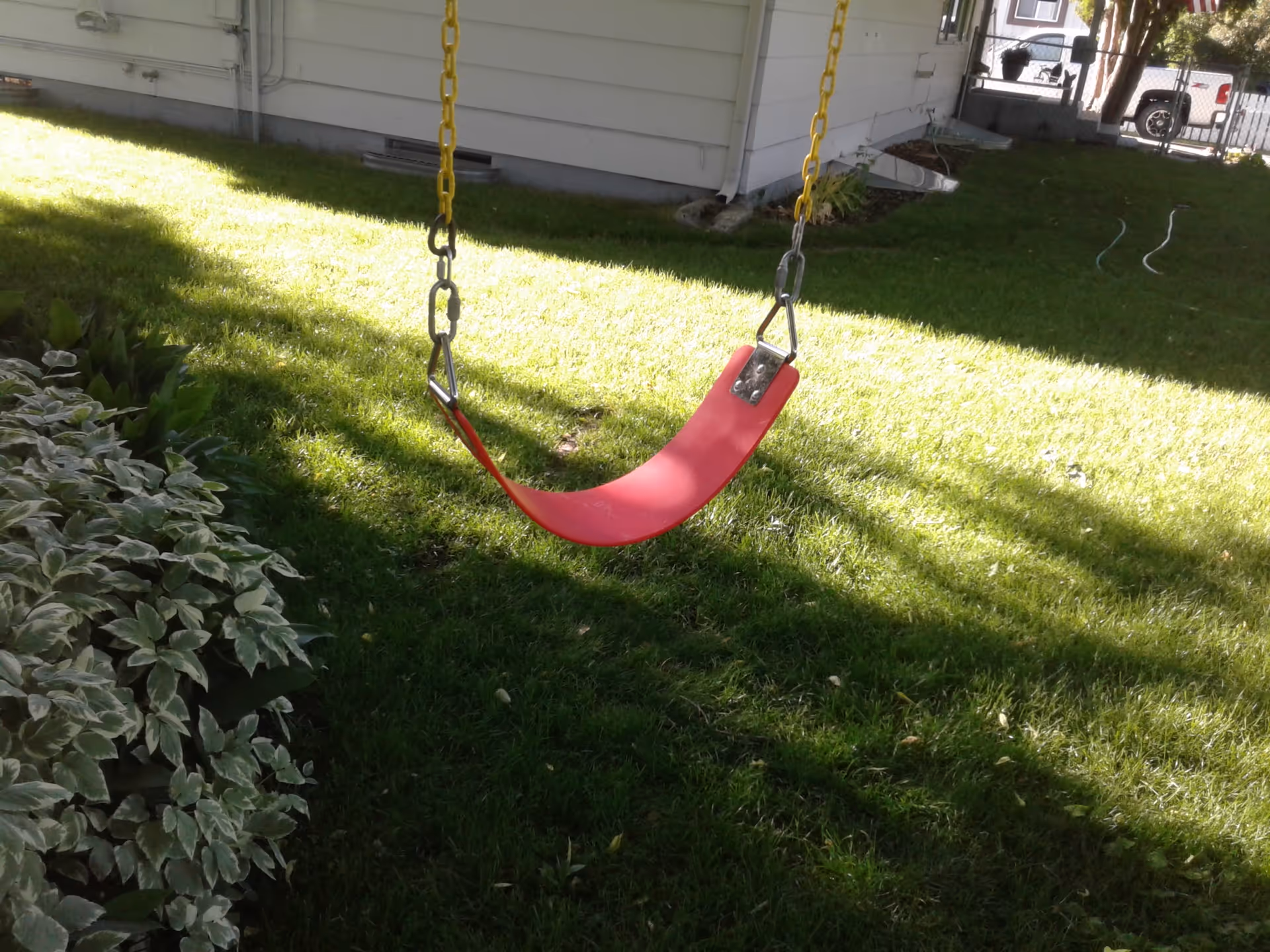 Red swing hangs from chains in a grassy yard beside a white house.