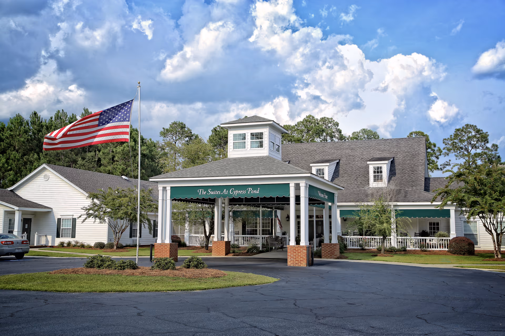 Front entrance of Cypress Pond Senior Care showing a covered porte-cochere, American flag, and landscaped grounds under a blue sky.