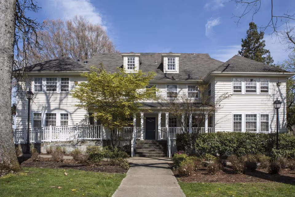 White two-story building with a front porch and surrounding trees and shrubs.
