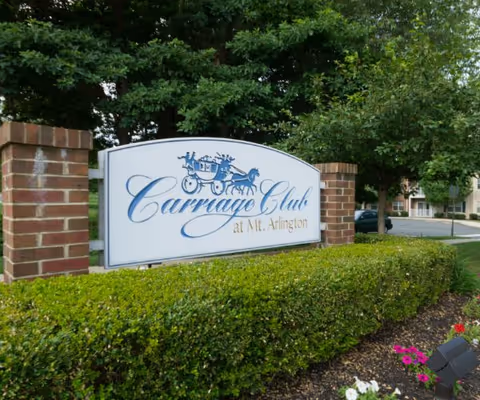 A Carriage Club sign mounted between brick pillars with greenery in a landscaped front yard.