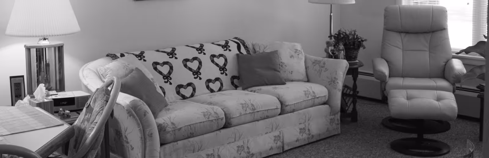 A cozy living room area featuring a floral-patterned sofa with a heart-patterned throw blanket and several cushions. To the right of the sofa is a cushioned swivel armchair. There is a side table with a lamp and a plant, and part of a dining table with chairs is visible on the left side of the image.