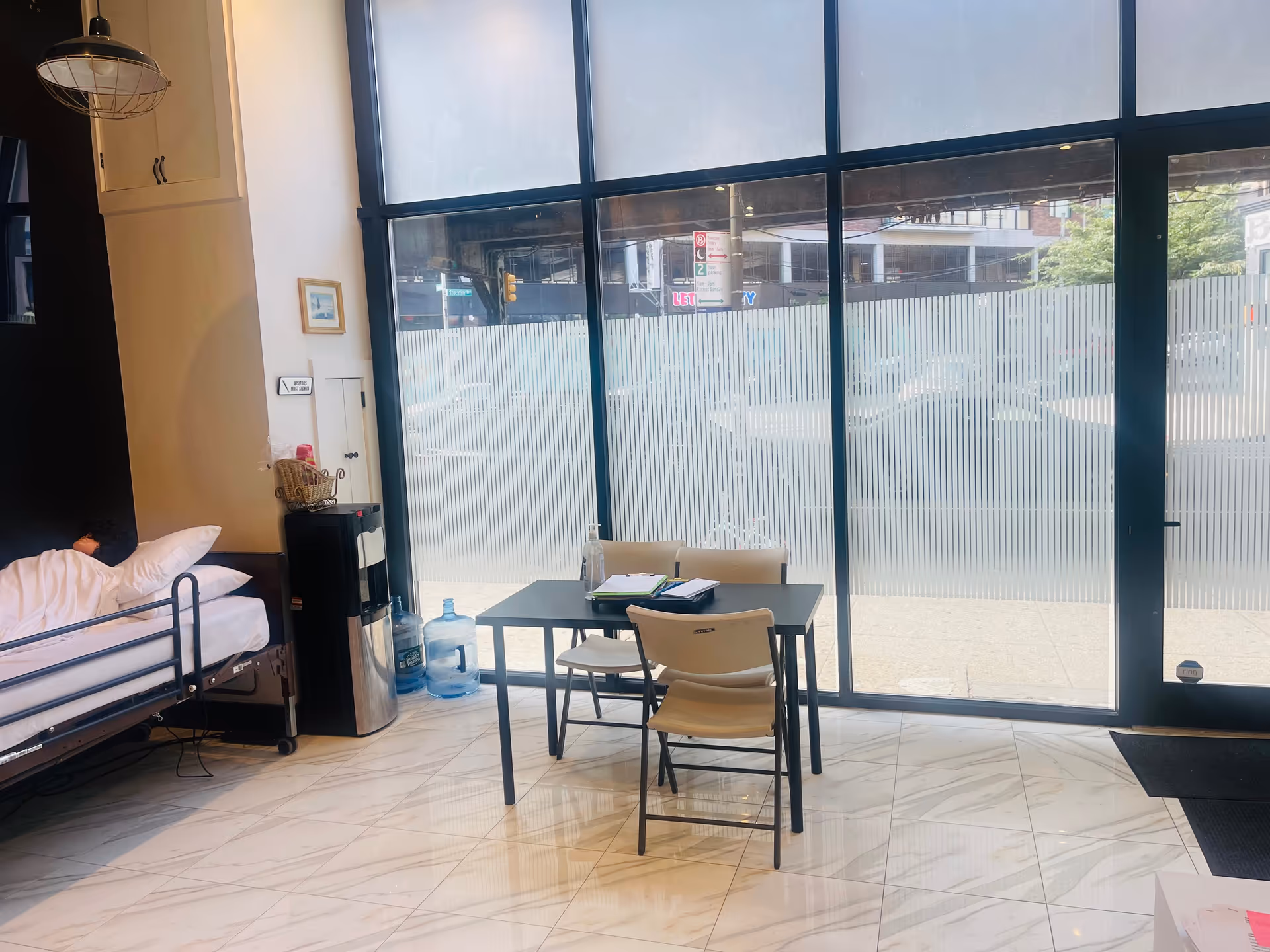 Bright interior with a hospital-style bed, table, and chairs by large glass doors.