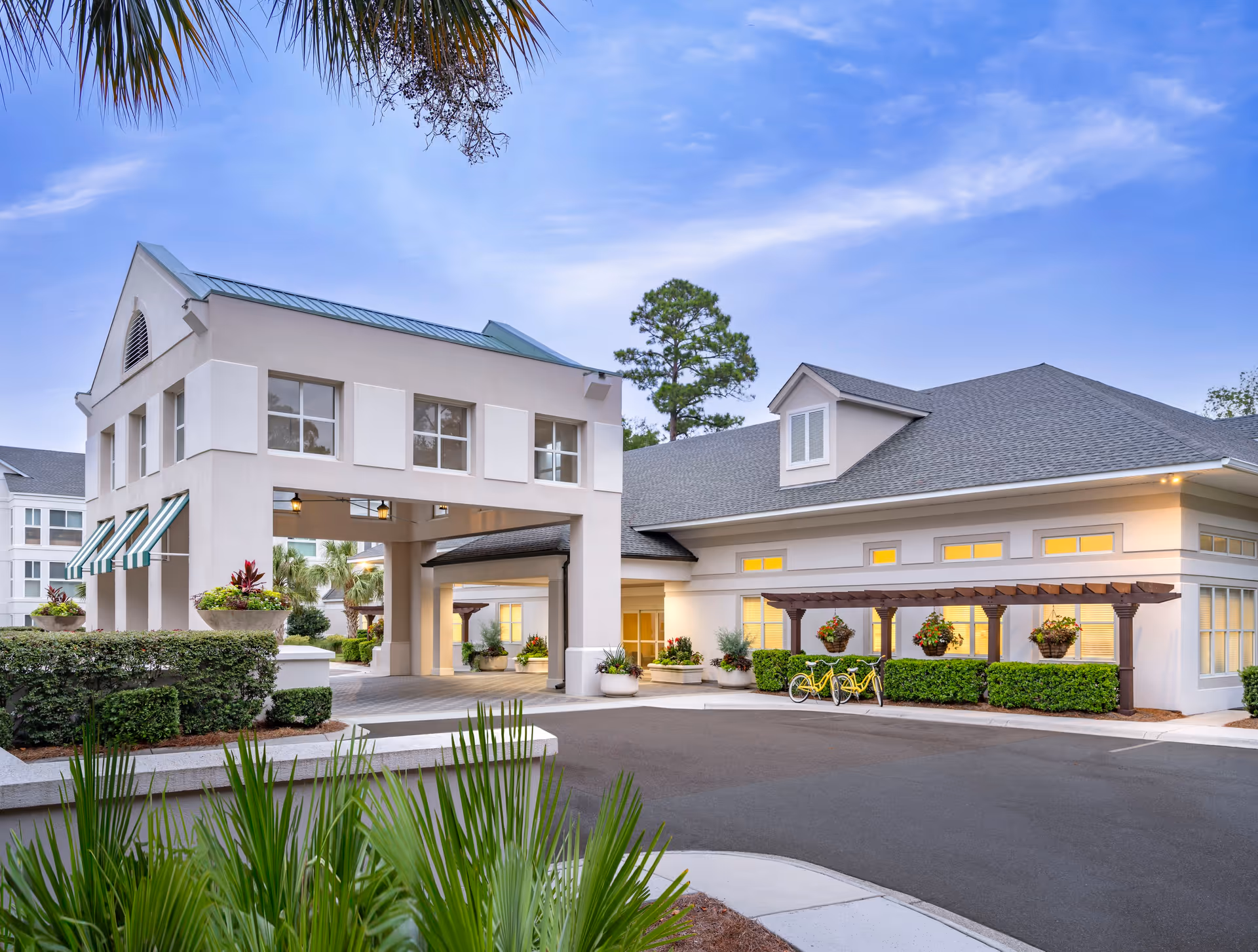 Exterior view of a senior living facility building with a covered driveway entrance, large windows, and a well-maintained landscape including bushes and palm trees under a clear blue sky.