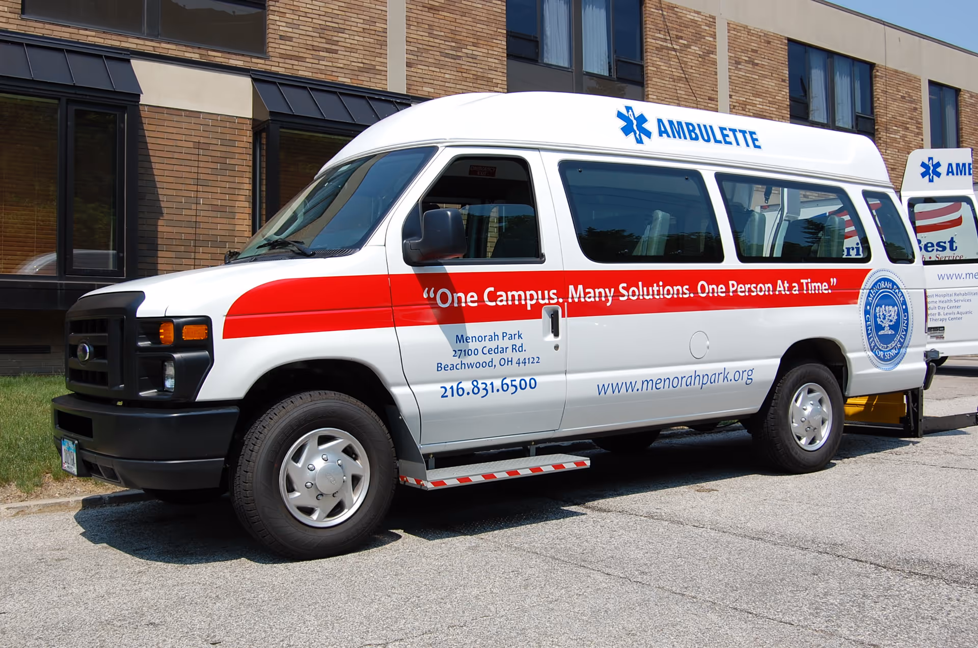 White ambulatory transport van with red stripe parked outside a brick Menorah Park building.