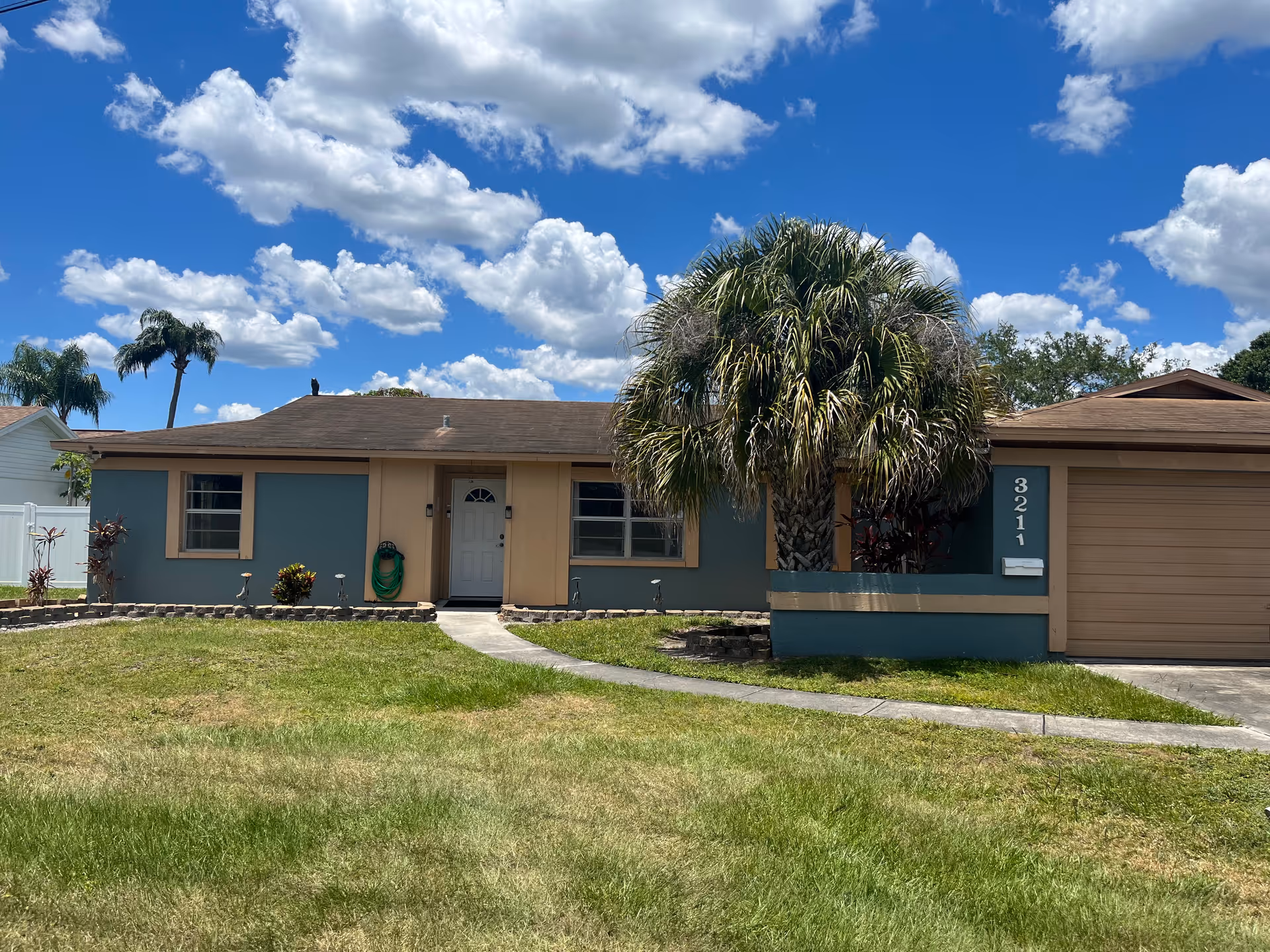 Single-story ranch-style house with a front lawn, large palm tree, and attached garage under a blue sky.