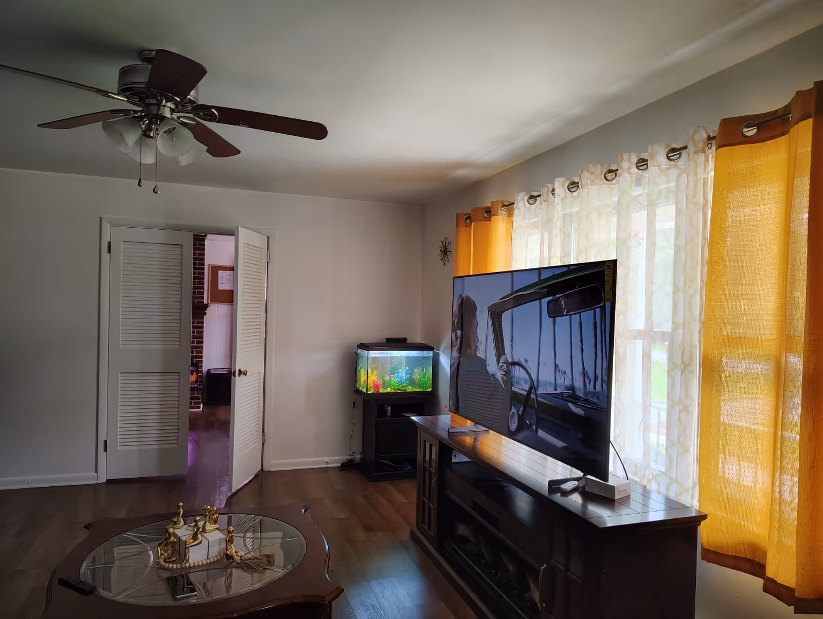 Living room with a large TV on a stand, aquarium, glass coffee table, and yellow curtains.