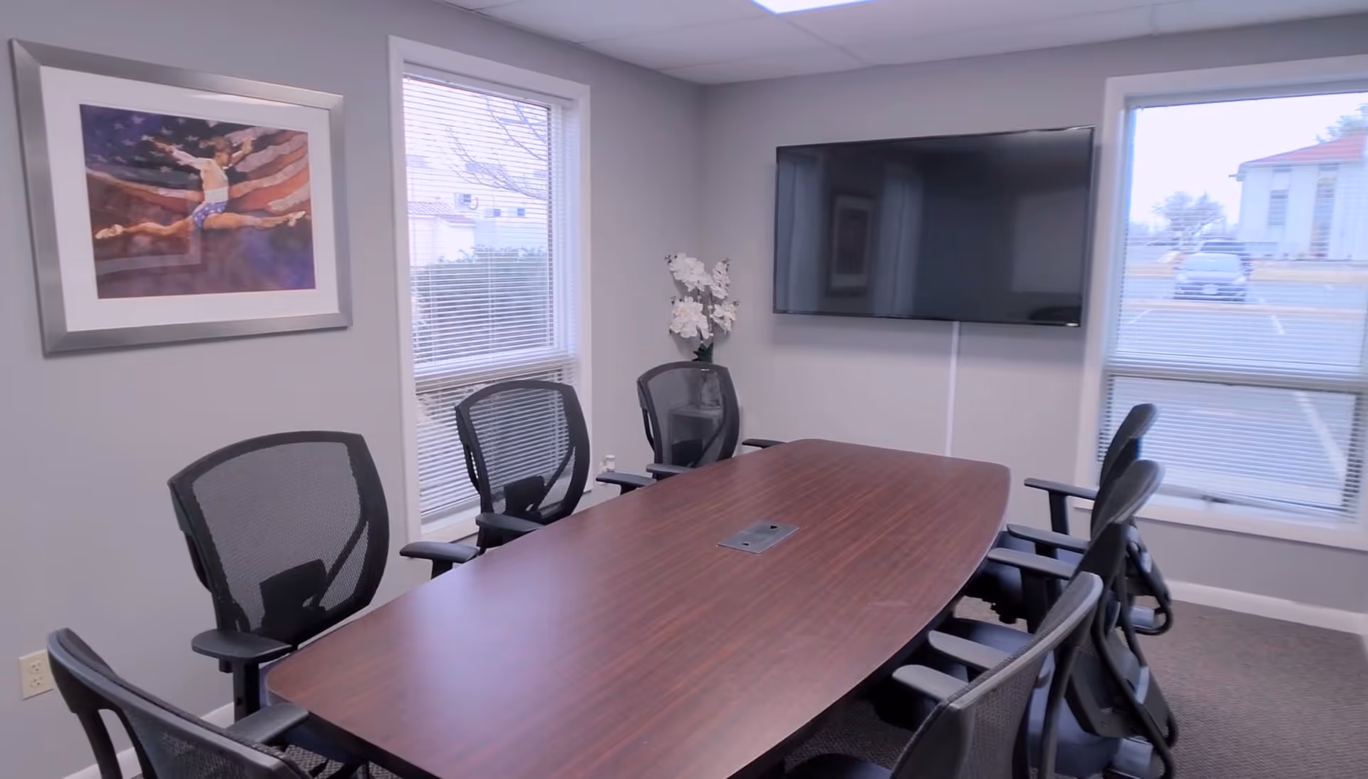 Conference room with a long wooden table, office chairs, wall-mounted TV, and windows.