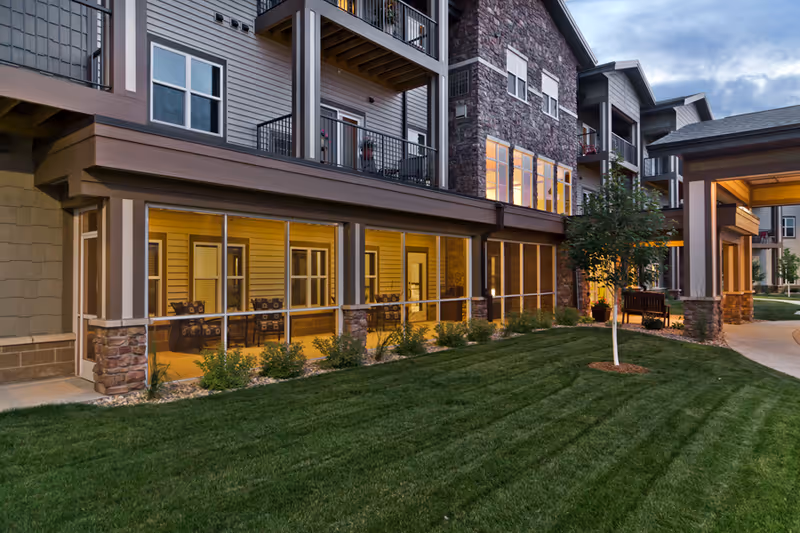 Exterior view of a multi-story senior living facility building at dusk with well-maintained green lawn, small trees, and a covered entrance. The building features a combination of stone and siding with balconies and large windows, some of which are lit from inside.