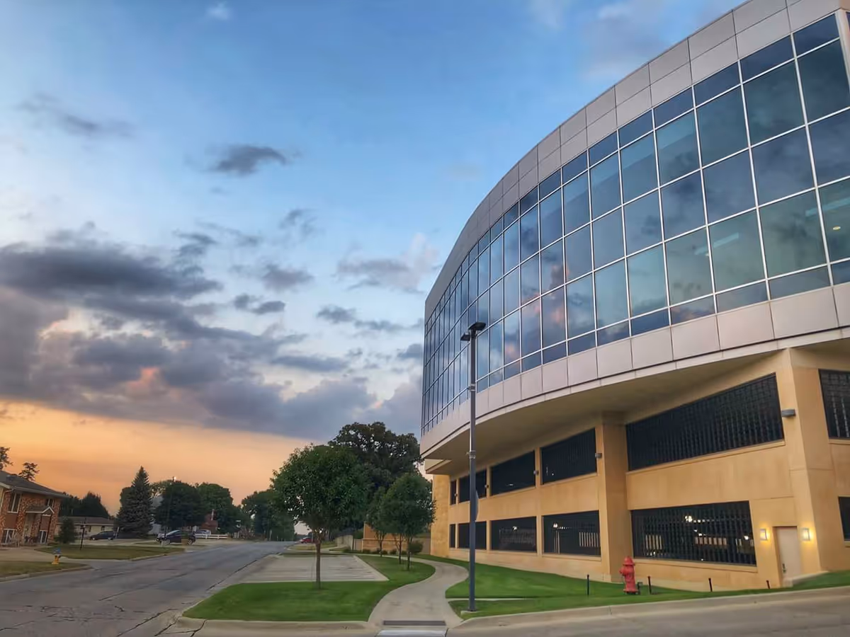Curved glass hospital exterior with sidewalk, trees, and a sunset sky.
