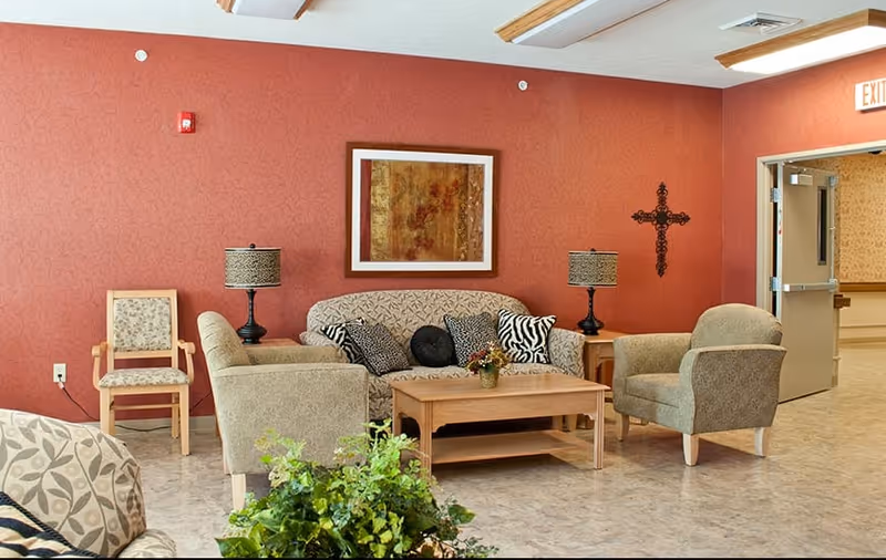 A cozy sitting area in a senior living facility with a patterned sofa, two armchairs, a wooden coffee table with a small plant, two table lamps with patterned shades, a framed abstract painting on a red wall, a decorative cross, and an open door leading to a hallway.