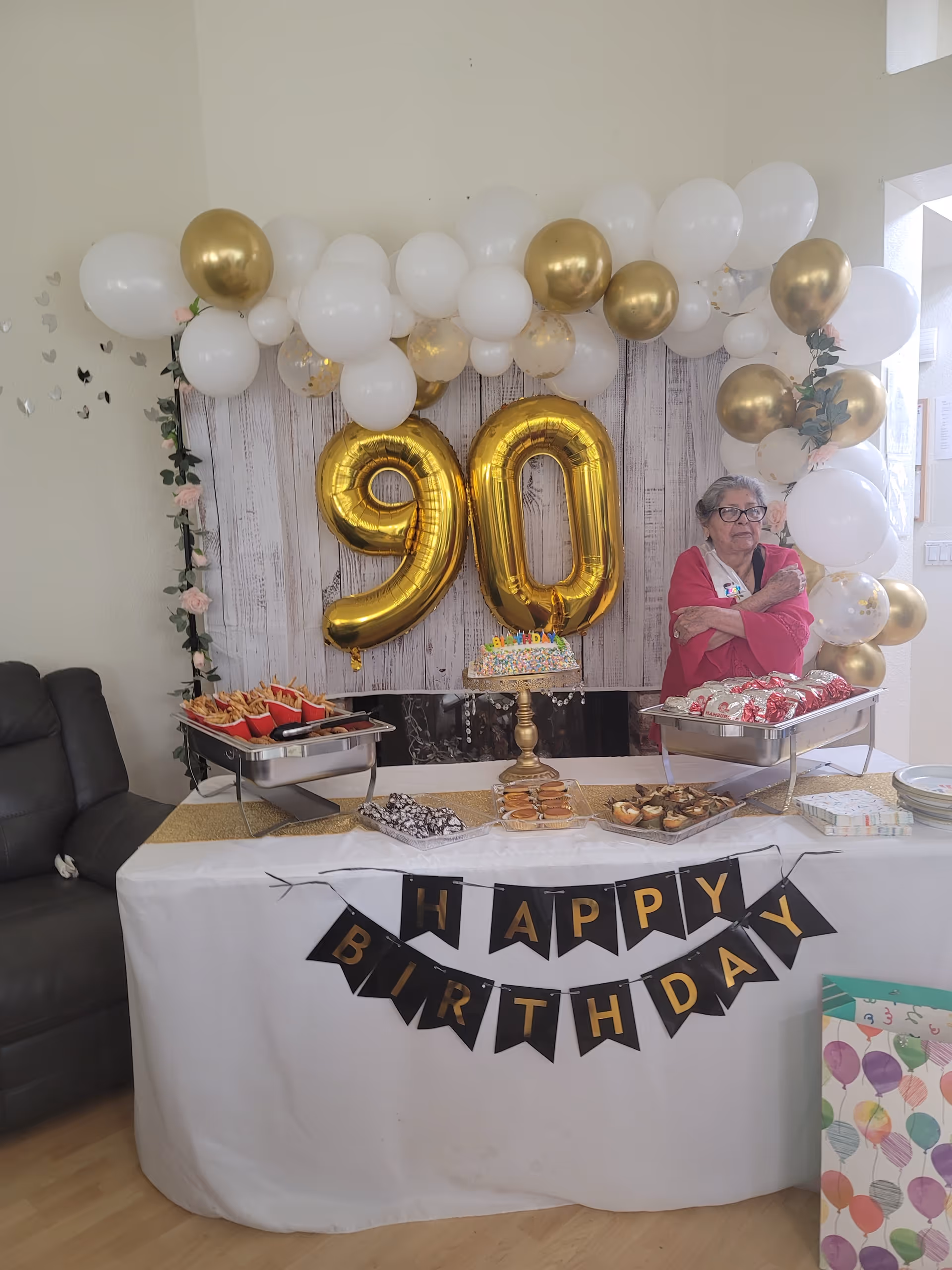 Elderly woman in pink stands beside a birthday table with gold '90' balloons and treats.