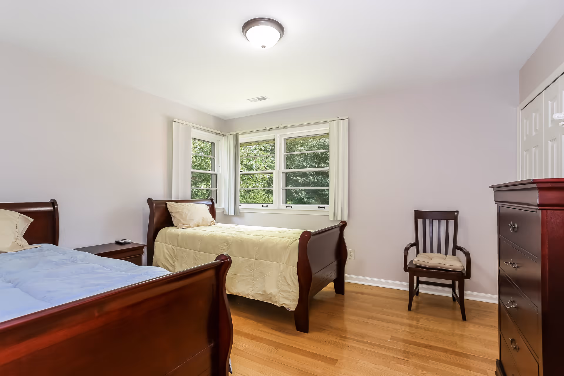 Two wooden single beds in a bright, neatly furnished bedroom with a window and dresser.