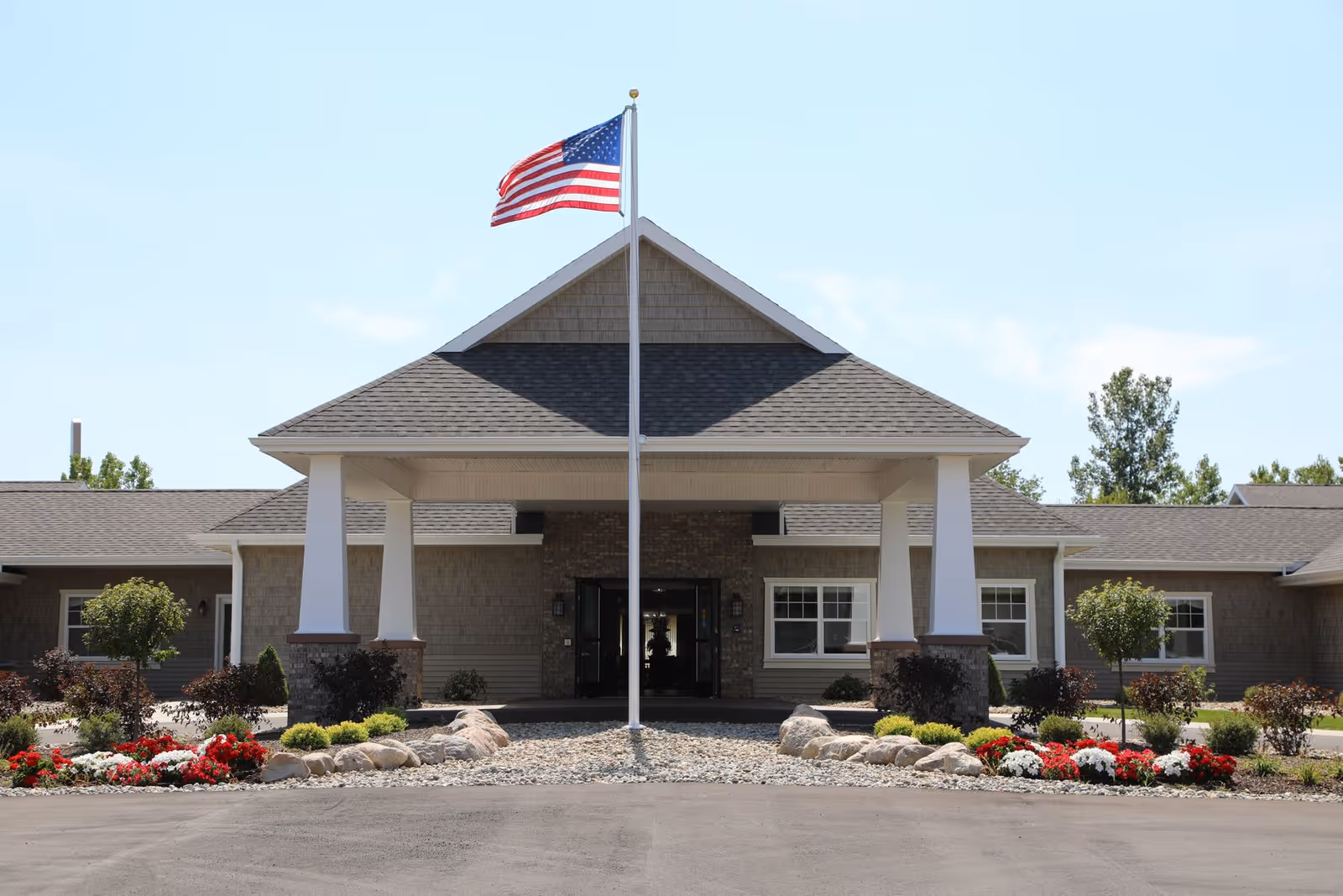 Front exterior view of The Brook of Portland facility with a covered entrance supported by white columns, an American flag on a flagpole in front, and landscaped flower beds with red and white flowers and small bushes.