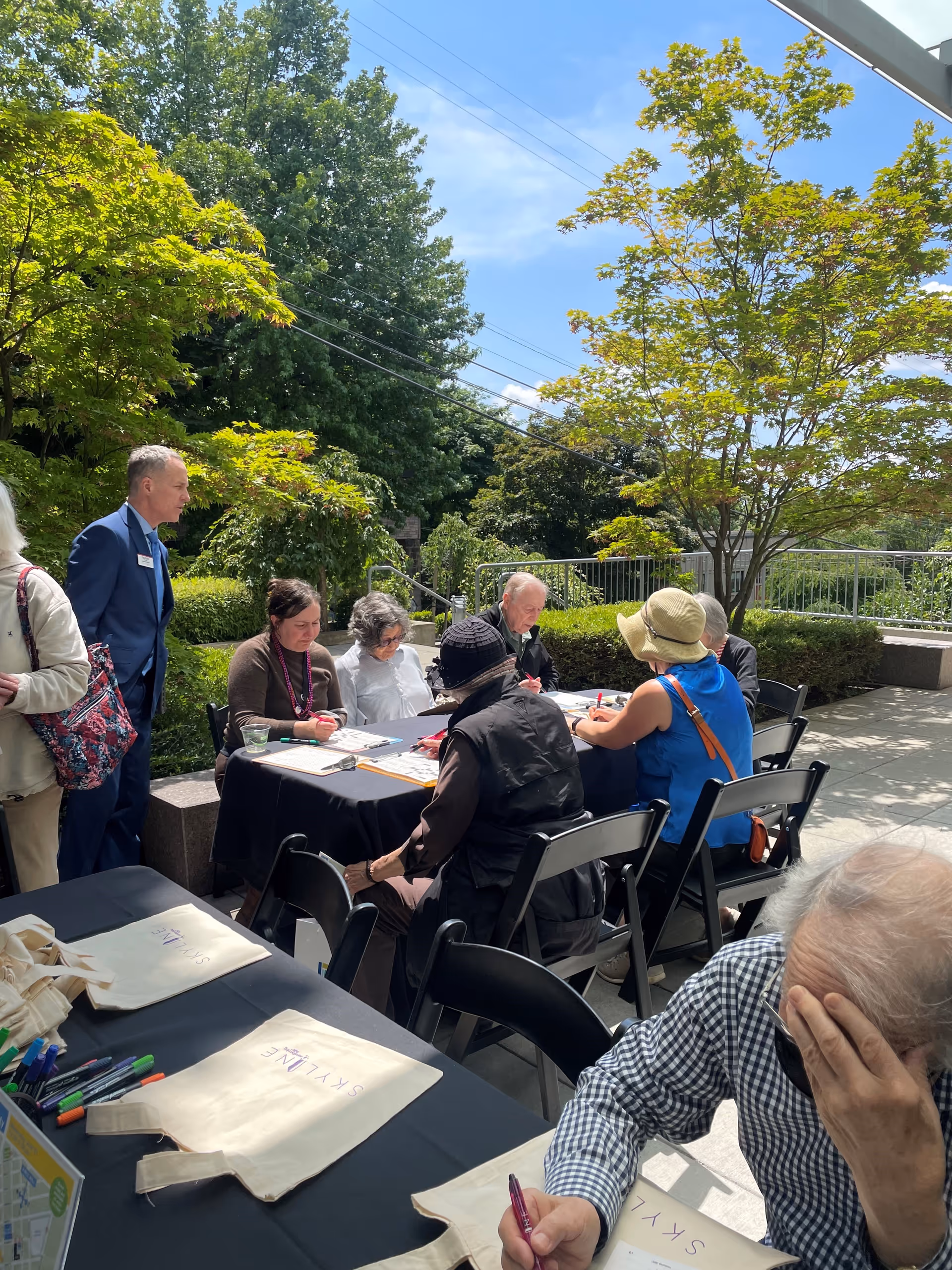 A group of elderly people sitting around a table outdoors on a sunny day, engaged in writing or drawing activities. A man in a blue suit stands nearby, and there are trees and greenery in the background. Tote bags with the word 'SKYLINE' are placed on a nearby table.
