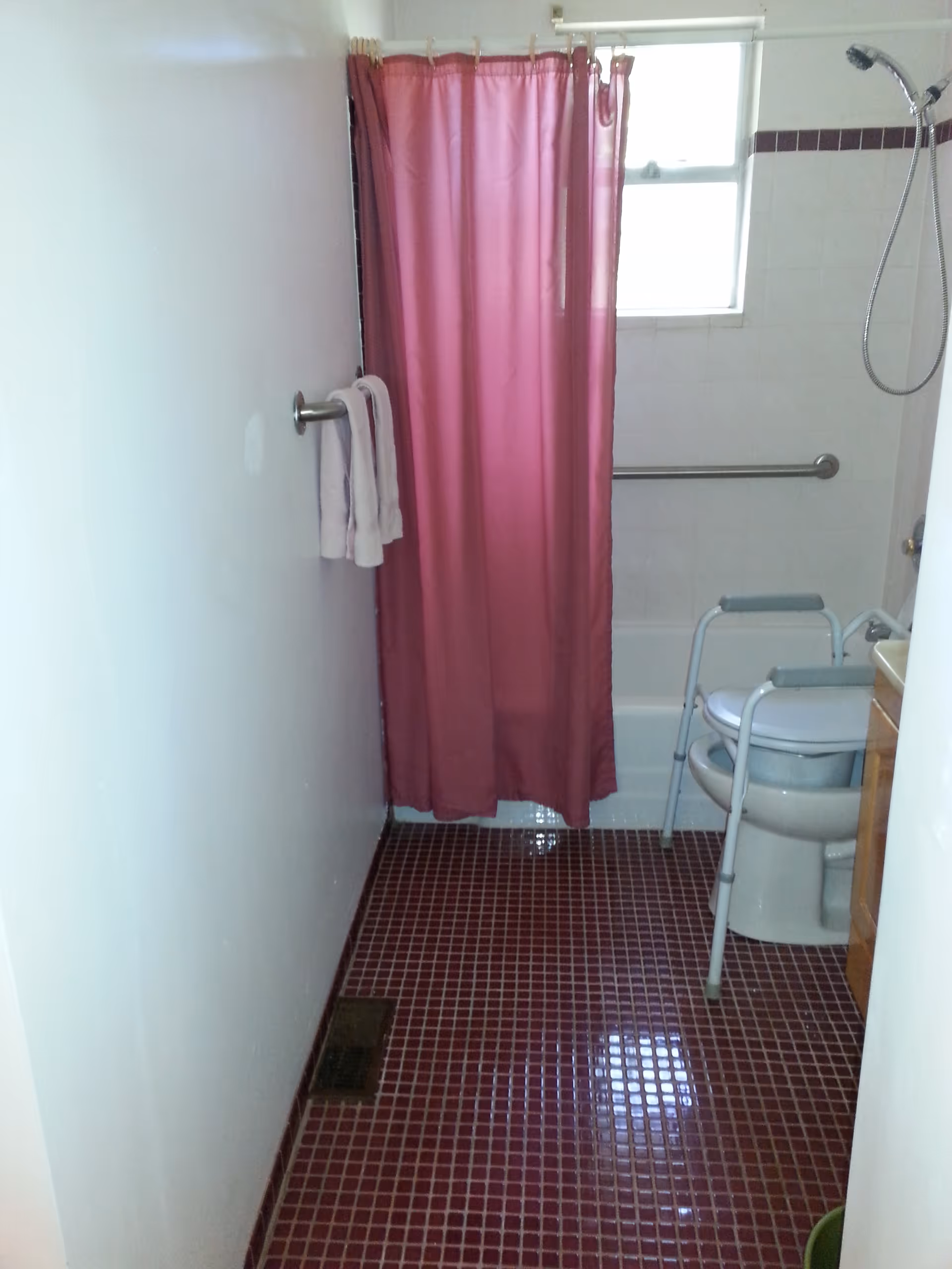 A small bathroom with a maroon tiled floor, a white wall with a towel rack holding two white towels, a bathtub with a pink shower curtain, a window above the tub, a handheld showerhead, a grab bar on the wall, and a toilet with a raised seat and armrests for accessibility.