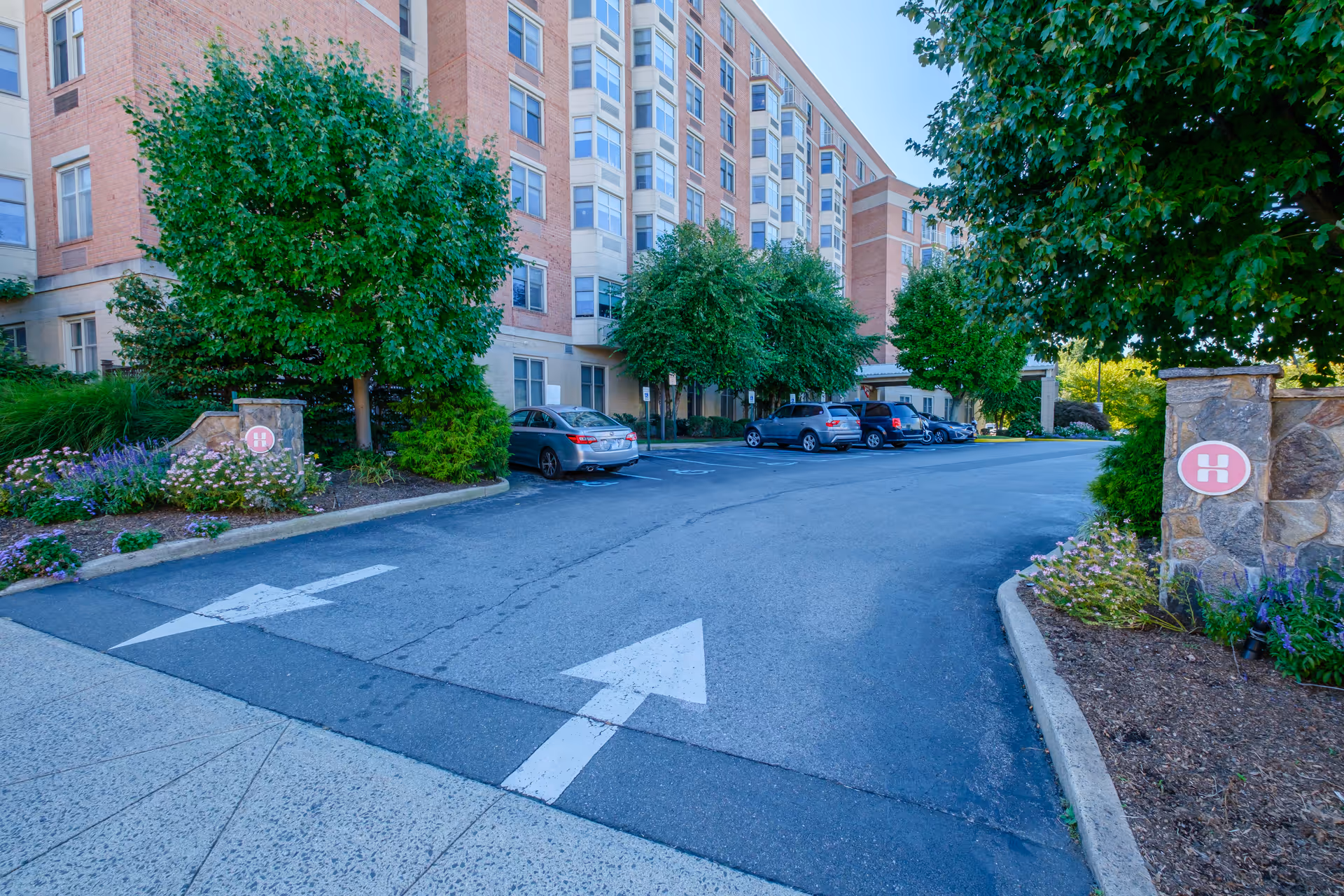Parking lot area outside a multi-story brick building with several cars parked along the side. The building has multiple windows and is surrounded by green trees and landscaped flower beds. There are white directional arrows painted on the asphalt indicating traffic flow.