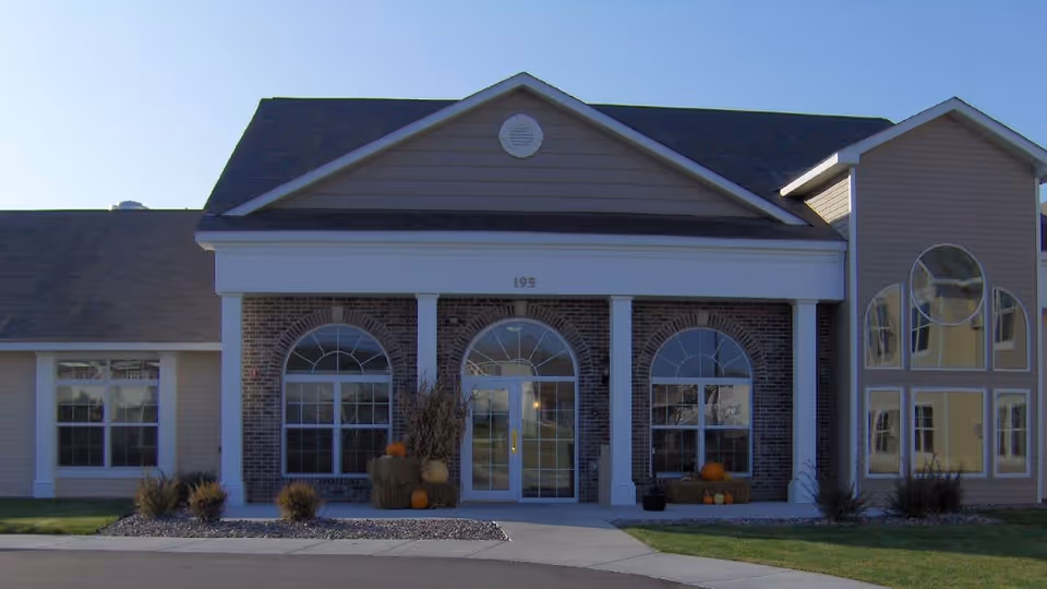 Front exterior view of a senior living facility building with a peaked roof, large arched windows, and a central entrance door. There are decorative pumpkins and hay bales near the entrance, and the building number 195 is visible above the door.