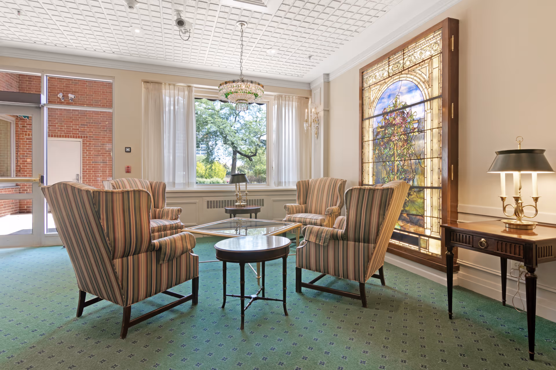 A bright seating lounge with four striped armchairs around a glass coffee table, a stained-glass window, chandelier, and large windows overlooking trees.