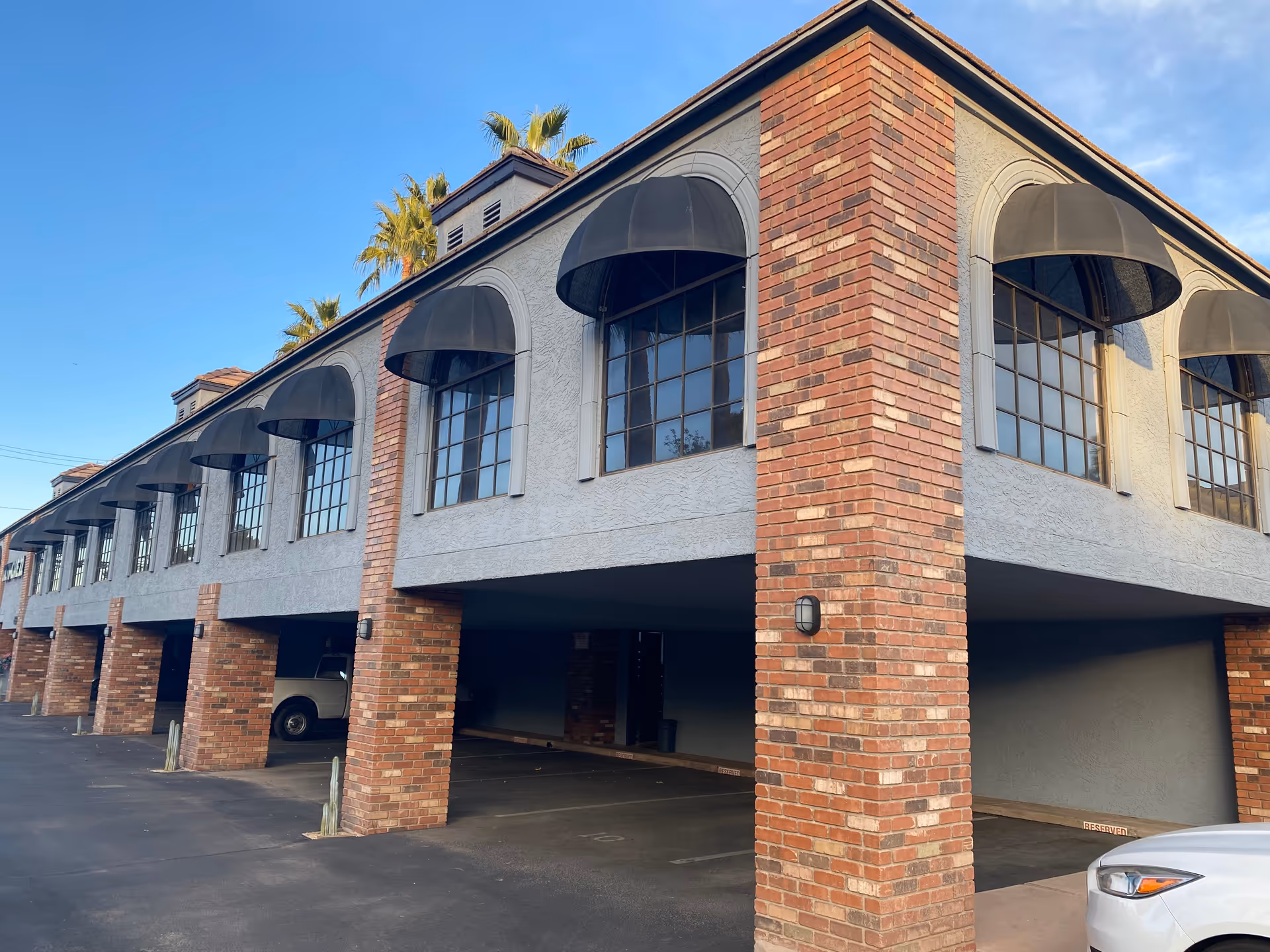 Two-story brick senior living building with arched windows and covered parking under a clear sky.