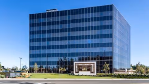 A modern glass-front office building with a landscaped entrance under blue sky.