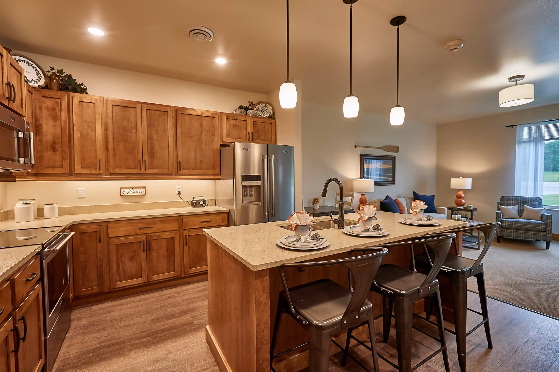 A modern kitchen with wooden cabinets, a stainless steel refrigerator, and a kitchen island with three metal bar stools. The island is set with plates, napkins, and cutlery. The kitchen opens into a living area with a window, armchair, and lamps providing warm lighting.