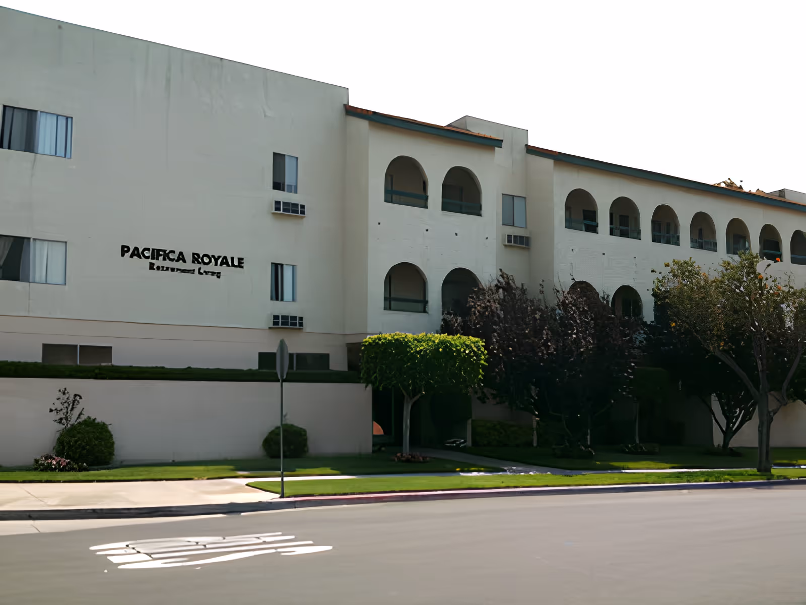 Exterior view of a multi-story residential building named Pacifica Royale with arched balconies, several windows, and neatly trimmed bushes and trees in front along a paved street.