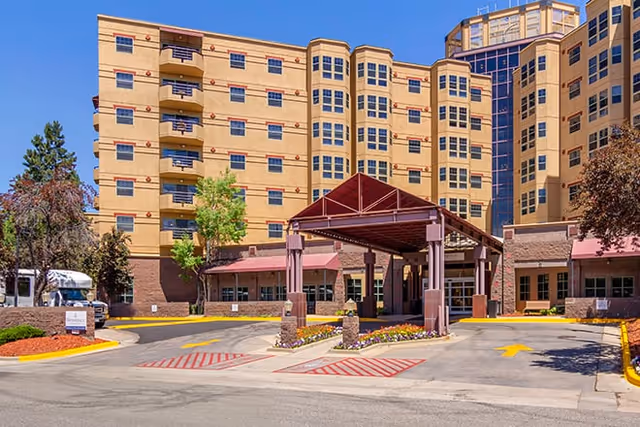 Front entrance and porte-cochère of a multi-story senior living building with landscaped driveway under a clear blue sky.