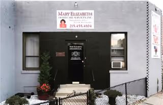 Exterior view of Mary Elizabeth Homecare Services entrance, black facade, steps, window, plants, and fence.