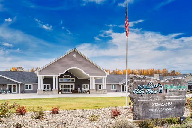 Front entrance of an assisted living facility with a covered porte-cochere, American flag, landscaped lawn, and a stone sign at the driveway.