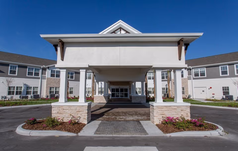 Front exterior view of The Crossings at Riverview facility showing a covered entrance with white pillars and stone bases, surrounded by a driveway and landscaped flower beds under a clear blue sky.