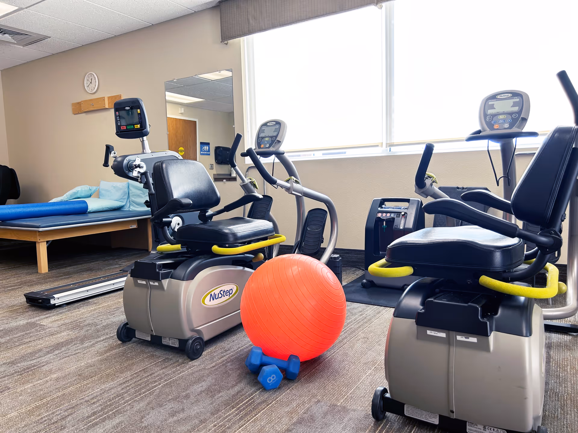 Rehabilitation gym with stationary bikes, a red exercise ball, dumbbells, and a bed in background.