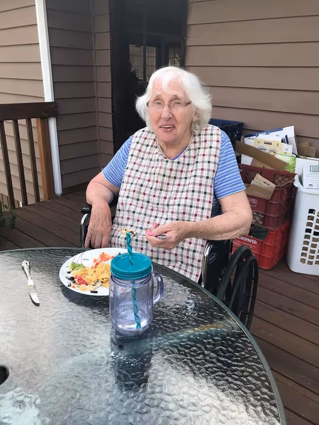 An elderly woman in a wheelchair sits at an outdoor glass table with a plate.