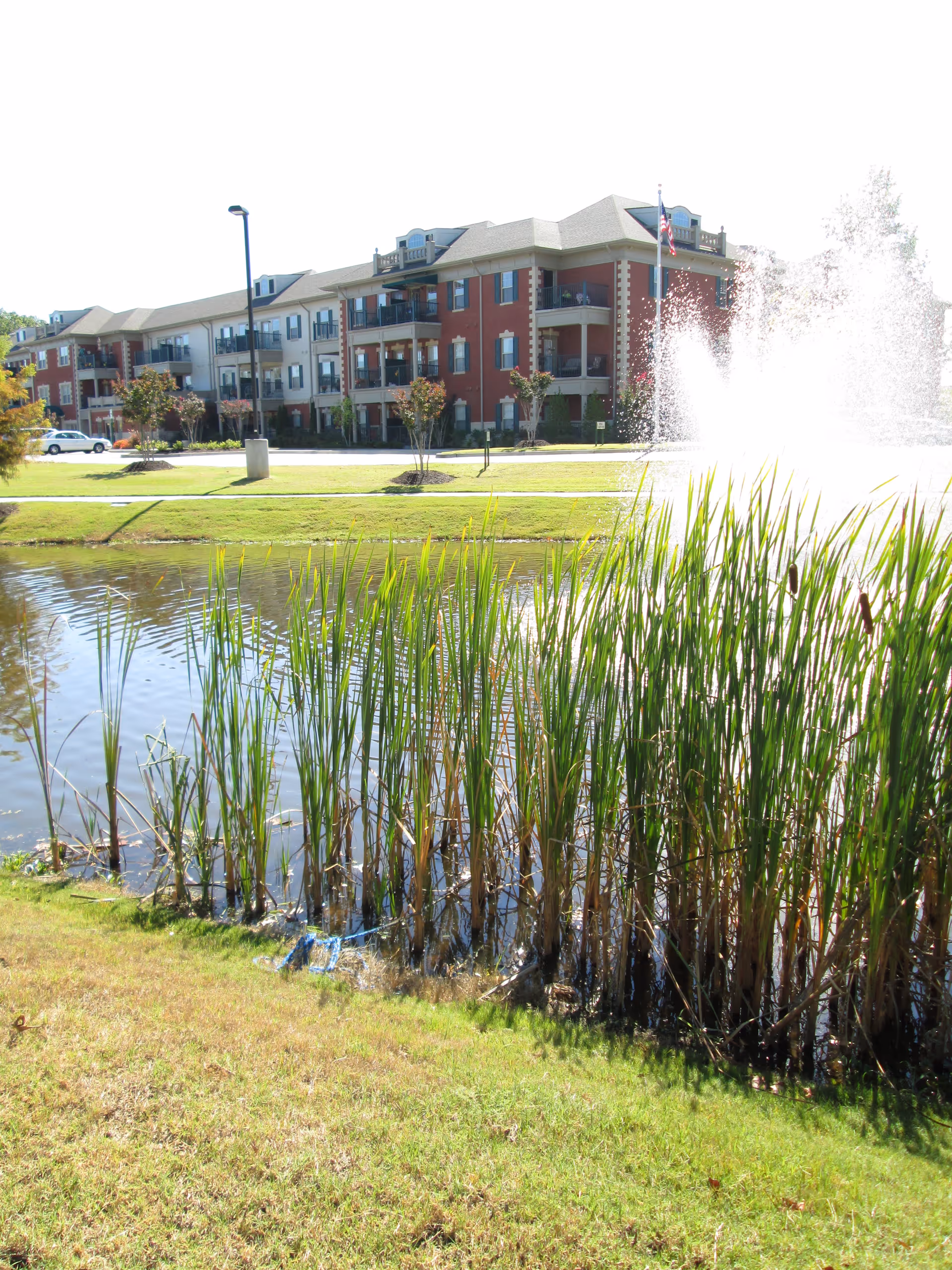 A senior living community building with three stories, featuring balconies and a mix of red brick and beige siding. In the foreground, there is a pond with tall green reeds and a water fountain spraying water high into the air. The area around the pond is grassy and well-maintained.